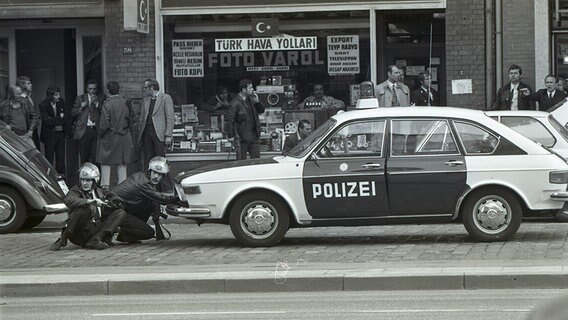 Polizisten verschanzen sich hinter einem Polizeiauto. Männer stehen vor einem Im-und Exportgeschäft. © Staatsarchiv Hamburg Plankammer (720-1_388_00_79877_18) 