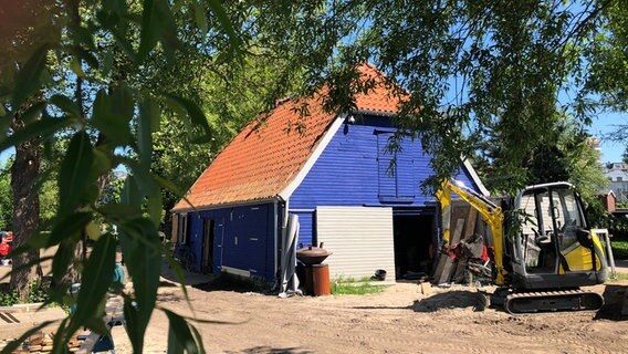 Ein blaues Haus mit roten Dachziegeln, im Vordergrund ein Bagger © NDR Foto: Nais Marie Baier